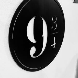 Könnte beinhalten: Ein schwarzes rundes Schild mit der Nummer "9 3/4", die aus der Mitte ausgeschnitten ist. Das Schild ist aus Metall und hat einen leicht erhöhten Rand.