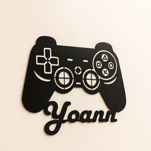 Puede incluir: Mando de videojuego de madera negra con detalles blancos. El mando presenta una cruceta, botones y el nombre "Yoann" en fuente cursiva. Un artículo decorativo para jugadores.
