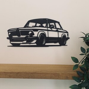 Peut inclure: Silhouette noire en métal d'une voiture classique, une BMW 2002, montée sur un mur au-dessus d'une étagère en bois.