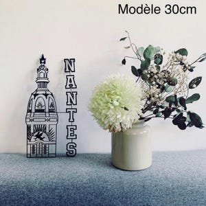 Peut inclure: Décoration murale en métal noir et blanc représentant un bâtiment et le mot "NANTES". Un vase avec des fleurs blanches et vertes est à droite. Le texte "Modèle 30cm" est en haut.
