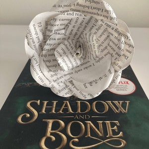Puede incluir: Una flor de papel blanco hecha con las p&aacute;ginas de un libro. La flor est&aacute; colocada encima de un libro titulado "Shadow and Bone" de Leigh Bardugo. La portada del libro es verde con letras doradas.