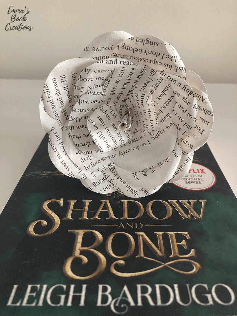 Puede incluir: Una flor de papel blanco hecha con las p&aacute;ginas de un libro. La flor est&aacute; colocada encima de un libro titulado "Shadow and Bone" de Leigh Bardugo. La portada del libro es verde con letras doradas.