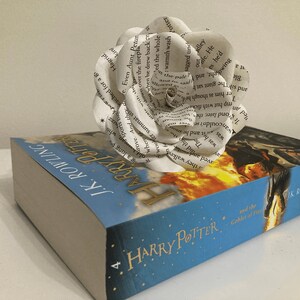 Puede incluir: Una flor de papel blanca hecha con las p&aacute;ginas de un libro se coloca encima de una copia de Harry Potter y el C&aacute;liz de Fuego. La portada del libro muestra un drag&oacute;n volando por el aire con fuego saliendo de su boca.