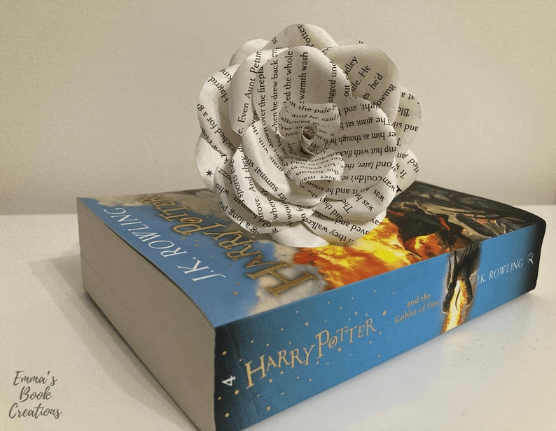 Puede incluir: Una flor de papel blanca hecha con las p&aacute;ginas de un libro se coloca encima de una copia de Harry Potter y el C&aacute;liz de Fuego. La portada del libro muestra un drag&oacute;n volando por el aire con fuego saliendo de su boca.