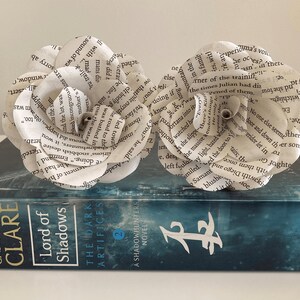 Puede incluir: Dos flores de papel blanco hechas con p&aacute;ginas de libros, colocadas sobre un libro azul titulado "Lord of Shadows" de Cassandra Clare. El libro es una novela de Cazadores de Sombras.