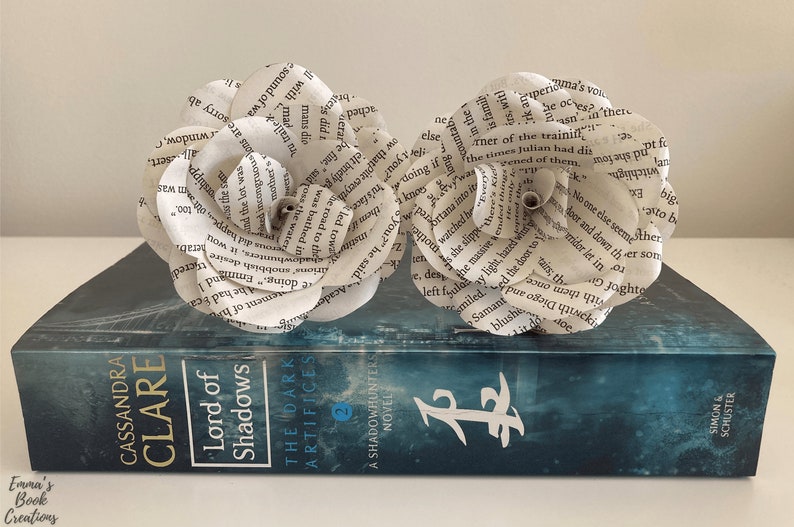 Puede incluir: Dos flores de papel blanco hechas con p&aacute;ginas de libros, colocadas sobre un libro azul titulado "Lord of Shadows" de Cassandra Clare. El libro es una novela de Cazadores de Sombras.