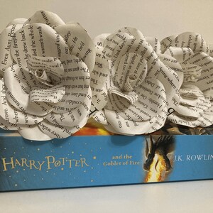 Puede incluir: Dos rosas blancas de papel hechas con p&aacute;ginas de libros est&aacute;n sobre un libro azul con un dise&ntilde;o de drag&oacute;n dorado. El libro se titula "Harry Potter y el C&aacute;liz de Fuego" de J.K. Rowling.