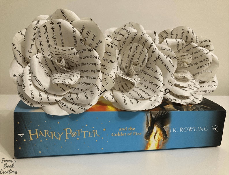 Puede incluir: Dos rosas blancas de papel hechas con p&aacute;ginas de libros est&aacute;n sobre un libro azul con un dise&ntilde;o de drag&oacute;n dorado. El libro se titula "Harry Potter y el C&aacute;liz de Fuego" de J.K. Rowling.