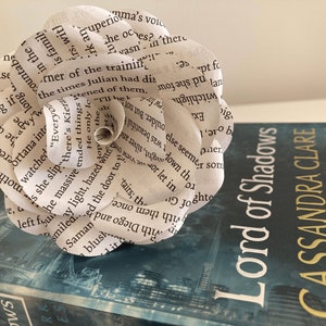 Puede incluir: Una flor de papel blanco hecha con las p&aacute;ginas de un libro, descansando sobre un libro azul titulado "Lord of Shadows" de Cassandra Clare. El libro forma parte de la serie "The Dark Artifices".