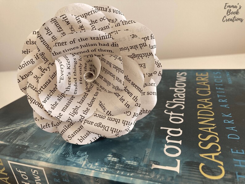 Puede incluir: Una flor de papel blanco hecha con las p&aacute;ginas de un libro, descansando sobre un libro azul titulado "Lord of Shadows" de Cassandra Clare. El libro forma parte de la serie "The Dark Artifices".
