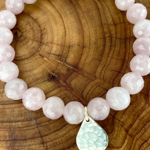 May include: A pink gemstone bracelet with a hammered silver teardrop charm. The bracelet is made of faceted beads and is strung on elastic.