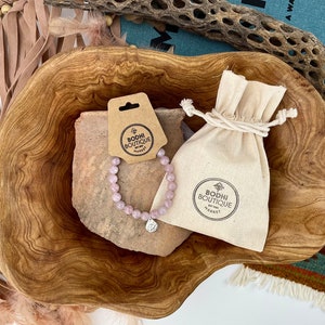 May include: A pink gemstone bracelet with a silver charm, displayed in a wooden bowl with a small white drawstring bag. The bag has a logo that reads "Bodhi Boutique by Sadh", and the tag attached to the bracelet reads "Bodhi Boutique".