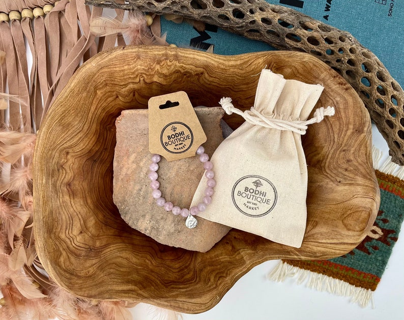 May include: A pink gemstone bracelet with a silver charm, displayed in a wooden bowl with a small white drawstring bag. The bag has a logo that reads "Bodhi Boutique by Sadh", and the tag attached to the bracelet reads "Bodhi Boutique".