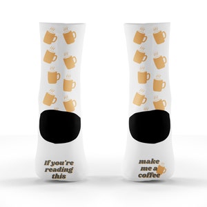 May include: A pair of white socks with a black toe and heel. The socks have a pattern of coffee cups with steam on them. One sock says "If you're reading this" and the other sock says "make me a coffee".