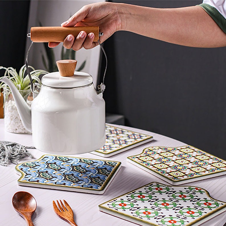 Puede incluir: Cuatro posavasos de cer&aacute;mica coloridos con patrones geom&eacute;tricos. Los posavasos tienen forma de rect&aacute;ngulo redondeado con una parte superior ligeramente puntiaguda. Una tetera de esmalte blanco con un asa de madera se est&aacute; levantando de uno de los posavasos.