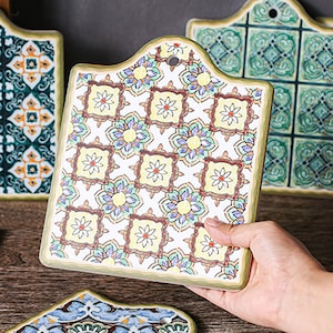 Puede incluir: Un juego de cuatro salvamanteles de cer&aacute;mica con un patr&oacute;n geom&eacute;trico colorido. Los salvamanteles tienen forma cuadrada con esquinas redondeadas y tienen un fondo blanco con acentos verdes, amarillos y marrones.