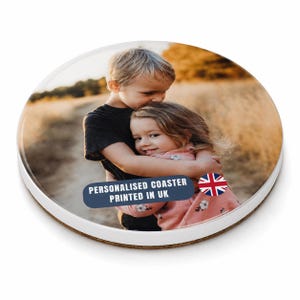 May include: A round ceramic coaster featuring a photograph of two children hugging in a field. A dark blue rectangular label on the coaster displays the white text "PERSONALISED COASTER PRINTED IN UK" next to a small circular Union Jack flag icon.