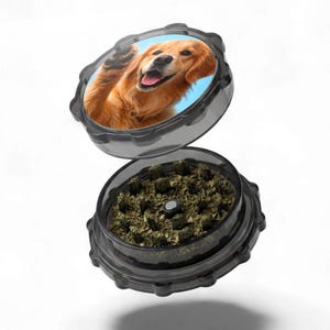 Puede incluir: Un molinillo de hierbas transparente de dos piezas con un exterior negro y estriado. La parte superior presenta un golden retriever sonriente contra un cielo azul. La sección inferior está llena de hierbas verdes molidas.