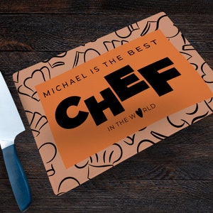 Pu&ograve; includere: Tagliere arancione con un motivo di cappelli da cuoco bianco e nero. Il tagliere ha un'etichetta rettangolare arancione con il testo "Michael &egrave; il miglior chef del mondo".