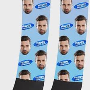 Puede incluir: Un par de calcetines azul claro con un patrón repetitivo del rostro de un hombre y gráficos ovalados azules que contienen el texto Happy Father's Day. Los calcetines tienen la puntera y el talón negros, con una banda negra en el puño superior.