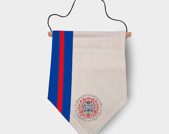 May include: A white fabric banner with a red and blue stripe on the left side. The banner has a royal blue circle with a crown and the text "Y Brenin Charles III Y Coroni 6 Mai 2023".