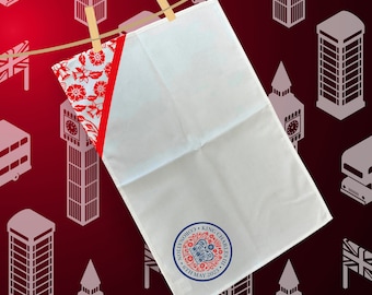 May include: White tea towel with a red and white floral design on one side and a royal blue and red coronation logo on the other side. The logo features a crown and the text "Coronation King Charles III 6th May 2023".