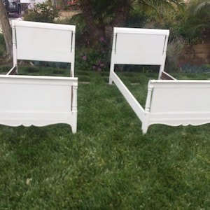 May include: Two white twin-sized bed frames with ornate headboards and footboards. The frames are on a grassy lawn.