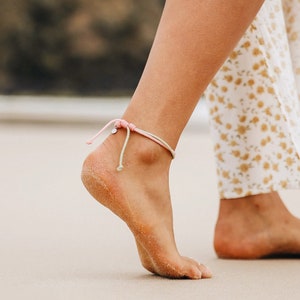 May include: A close-up of a person's ankle wearing a pink and green ankle bracelet with a silver charm. The bracelet is tied in a knot and the charm is a small circle.