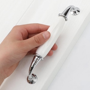 May include: A white ceramic cabinet handle with a smooth, elongated center and polished chrome metal ends. The chrome hardware features a curved design with circular mounting bases and the engraved text "Khan Malik" visible on the lower metal arm.