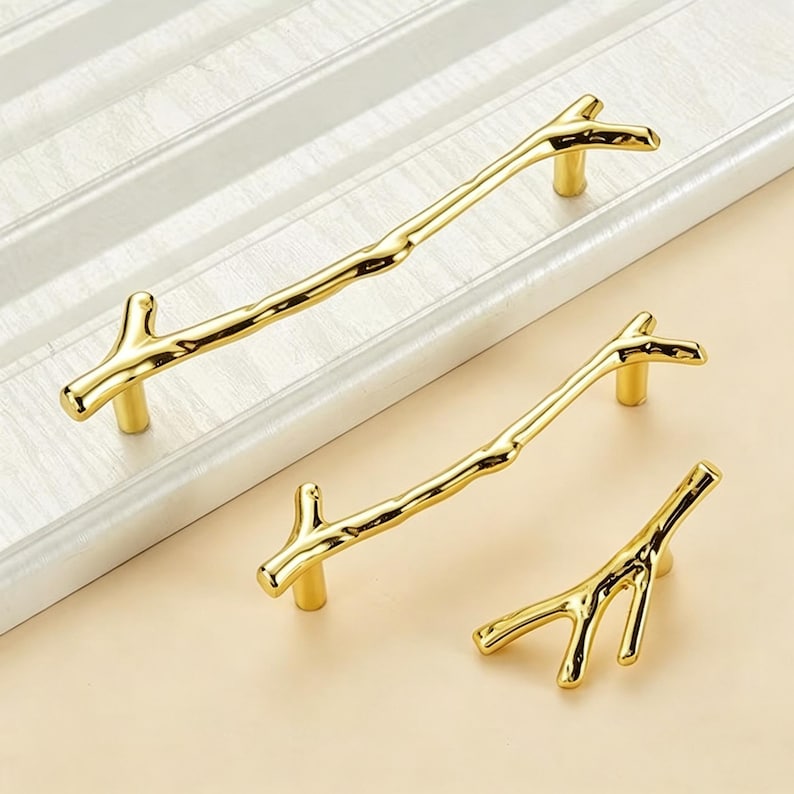 May include: Three gold-colored cabinet pulls shaped like tree branches. The set includes one long handle, one medium handle, and one small knob, all featuring a smooth, reflective metallic finish and a realistic twig-like texture with small offshoots.