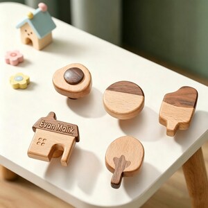 Puede incluir: Una colección de pomos de madera para cajones en varias formas, incluyendo una casa con el nombre Evan Malik grabado, un círculo, un óvalo irregular y un árbol. Los pomos presentan una combinación de tonos de madera clara y oscura.