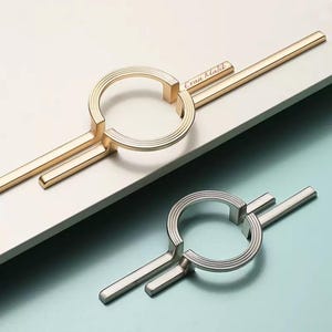 May include: Two metal cabinet pulls, one gold and one silver, featuring a circular centre with concentric grooved lines and two horizontal bars extending from the sides. The gold pull has the name Evan Malik engraved on the top horizontal bar.