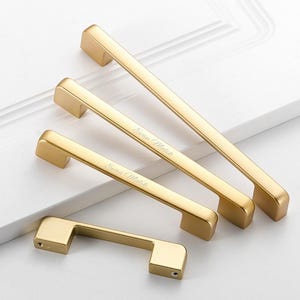 May include: Four gold-toned metal cabinet pulls of varying lengths arranged diagonally. Two of the pulls feature the engraved text "Evan Malik" in a cursive script. Each handle has a rectangular bar design with two mounting points at the ends.