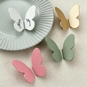Può includere: Quattro pomelli per armadietti a forma di farfalla in bianco, oro, verde salvia e rosa. Il pomello bianco presenta la silhouette di una persona su ogni ala. Ogni pomello è composto da due pezzi separati a forma di ala da montare insieme.