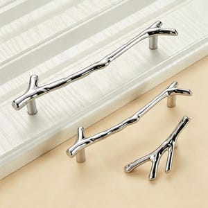 May include: Three silver-toned cabinet pulls shaped like tree branches with a smooth, reflective metallic finish. Each handle features a distinct, irregular twig-like structure with small offshoots, mounted on cylindrical posts for attachment.