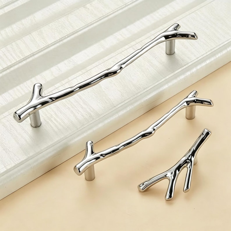 May include: Three silver-toned cabinet pulls shaped like tree branches with a smooth, reflective metallic finish. Each handle features a distinct, irregular twig-like structure with small offshoots, mounted on cylindrical posts for attachment.