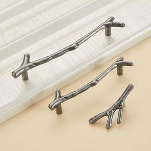 May include: Three cabinet pulls in a dark metallic finish, each shaped like a textured tree branch. The set includes two long horizontal handles and one smaller, Y-shaped knob, all featuring a rugged, organic appearance with visible knots.