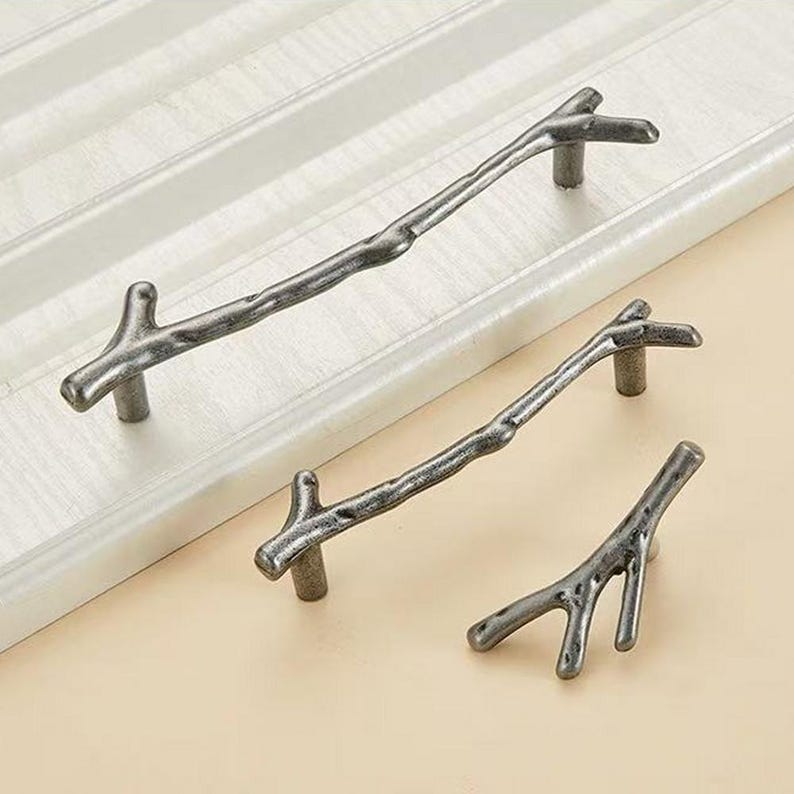 May include: Three cabinet pulls in a dark metallic finish, each shaped like a textured tree branch. The set includes two long horizontal handles and one smaller, Y-shaped knob, all featuring a rugged, organic appearance with visible knots.