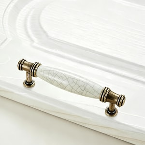 May include: A cabinet handle featuring a central white ceramic bar with a dark crackle pattern, flanked by two antique brass-toned metal ends with ribbed detailing. The handle is mounted horizontally on a white surface with a wood grain texture.