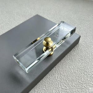 May include: A rectangular clear acrylic cabinet handle featuring a brass-toned metal base and a laser-etched graphic of a rabbit head on the top surface. The handle has beveled edges and a polished finish, providing a transparent look.
