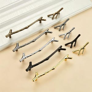May include: A collection of cabinet handles and knobs shaped like tree branches, available in various metallic finishes including antique bronze, brushed silver, polished chrome, matte black, and bright gold, arranged on a flat surface.