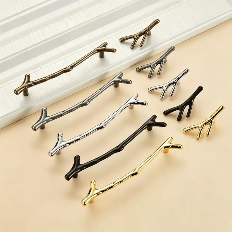 May include: A collection of cabinet handles and knobs shaped like tree branches, available in various metallic finishes including antique bronze, brushed silver, polished chrome, matte black, and bright gold, arranged on a flat surface.