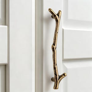 May include: A vertical cabinet handle shaped like a textured tree branch with a dark antique brass finish. The handle features a gnarled, organic appearance with small protruding twigs at the top and bottom, mounted onto a white cabinet door.