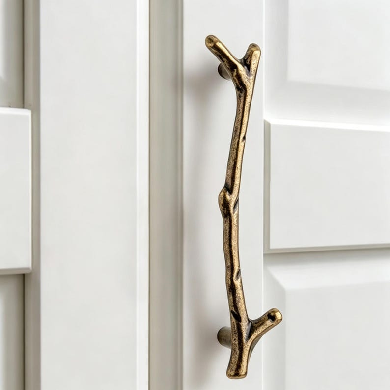 May include: A vertical cabinet handle shaped like a textured tree branch with a dark antique brass finish. The handle features a gnarled, organic appearance with small protruding twigs at the top and bottom, mounted onto a white cabinet door.