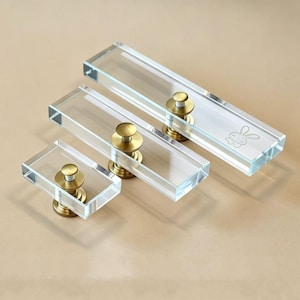Op de afbeelding: Drie rechthoekige ladeknoppen van helder acryl in verschillende lengtes, elk voorzien van een centrale messingkleurige metalen bevestigingsstijl. De grootste knop heeft een kleine, gegraveerde lijntekening van een konijnengezicht.