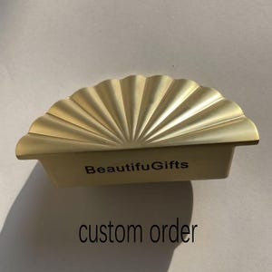 Peut inclure: Un plateau décoratif doré en forme d'éventail avec le texte "BeautifuGifts" et "custom order". Le plateau a un design festonné et une base rectangulaire. Le plateau est en métal et est probablement utilisé à des fins décoratives.