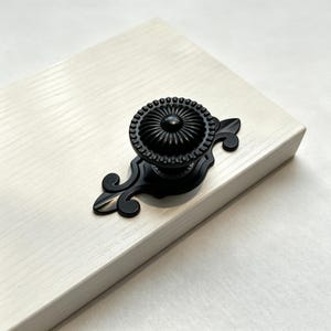 Puede incluir: Un pomo de mueble negro mate con una parte superior circular estriada y una cúpula central, montado sobre una placa trasera ornamentada y alargada con extremos curvos en forma de voluta. El herraje está fijado a un panel de madera clara.