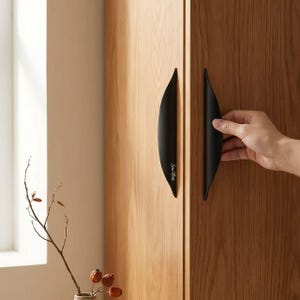 May include: Two matte black, crescent-shaped cabinet handles mounted vertically on a light brown wood-grain cabinet door. The handle on the right features the white script text "Sine Melle" printed along its curved edge, showcasing a sleek design.