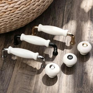 May include: Three cabinet pulls with white ceramic handles and metal bases in gold, black, and silver finishes, alongside three round ceramic knobs with gold, black, and silver accents. One pull features the text "Sine Melle" and one knob has "EM".