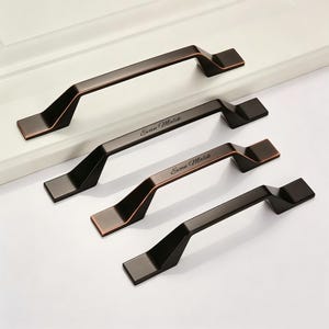 May include: Four dark bronze cabinet pulls with angled ends and a smooth finish. Two of the pulls feature the engraved text Evan Malik in a cursive script on the top surface. The hardware is arranged in a diagonal row on a white surface.